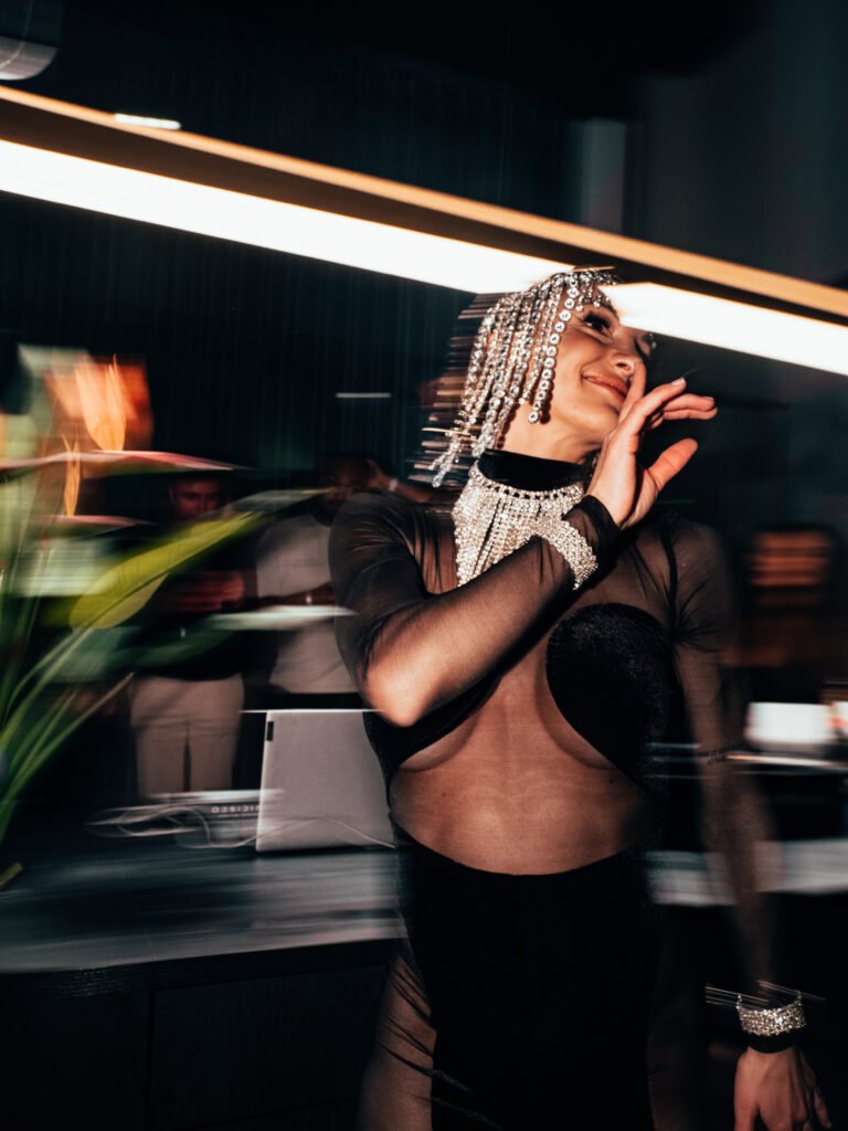 De foto toont een performer tijdens CB Nights, gekleed in een opvallende, elegante outfit met transparante details en glinsterende accessoires. De persoon draagt een opvallende hoofdtooi met glinsterende strengen en een bijpassende statement-halsketting. Door de beweging in de scène ontstaat motion blur, wat een dynamisch en energiek effect geeft. De achtergrond bevat zachte verlichting en vage silhouetten van gasten, waardoor de performer centraal en krachtig naar voren komt.