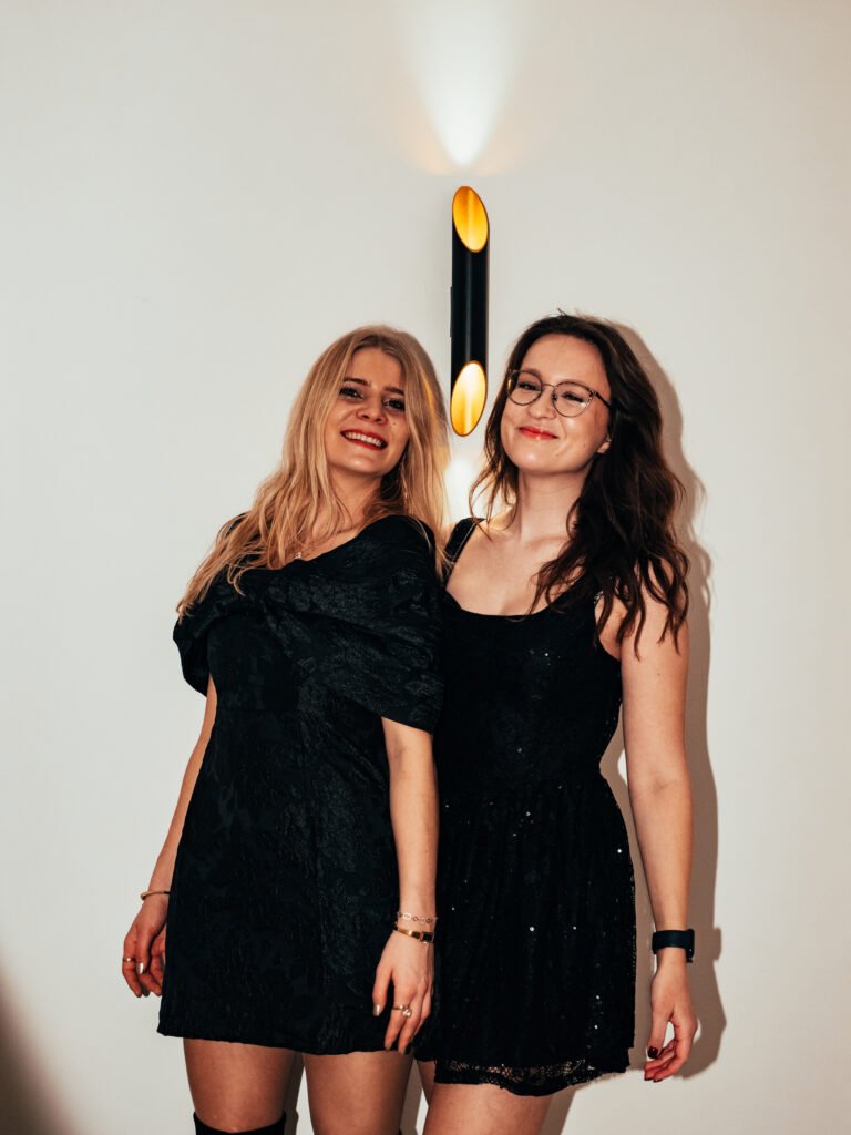 Marie Hendrikx in zwarte feestjurken die samen poseren voor een witte muur met wandlamp.