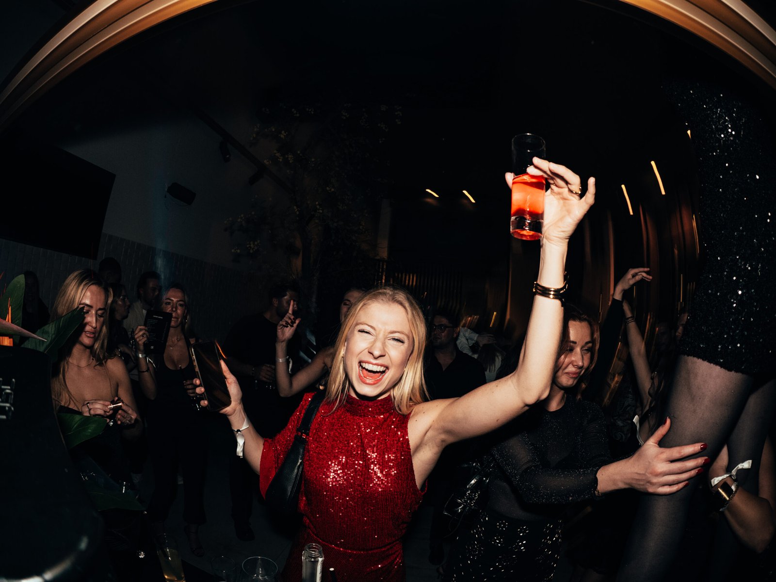 De foto toont een energieke en feestelijke scène tijdens CB Nights. Centraal staat een vrouw in een opvallende rode glitterjurk die lacht en een glas met rood drankje omhoog houdt. Rondom haar dansen andere gasten, terwijl warme belichting en lichte motion blur de dynamiek en intensiteit van de avond benadrukken. De sfeer is druk, luxe en vol beleving.